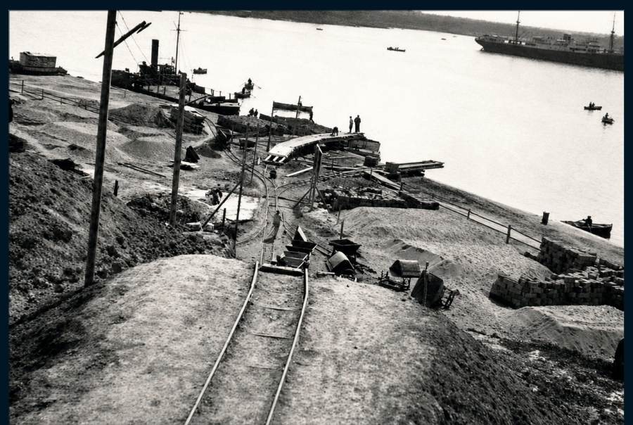 Die Vorbereitung der Baustelle: die Schienen für den Materialtransport; im Hintergrund ein Schiff im Hafen.